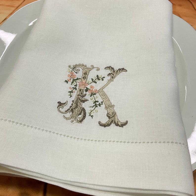 May include: White linen napkin with a brown embroidered letter K surrounded by small pink flowers.
