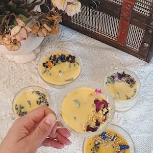 May include: Five small glass candle jars with yellow wax and dried flowers. The candles are arranged on a white lace tablecloth.