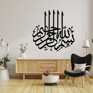 May include: A black and white wall decal with Arabic calligraphy that reads "Bismillah" in a decorative font. The decal is displayed on a white wall in a living room setting with a wooden cabinet, a chair, and plants.