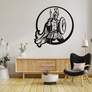 Puede incluir: Silueta en blanco y negro de un guerrero sosteniendo una lanza y un escudo en un marco circular. El guerrero lleva un casco y una armadura. La imagen est&aacute; en una pared blanca con un armario de madera y una silla en el fondo.