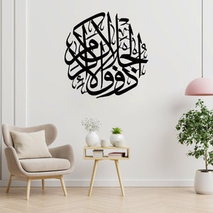 Op de afbeelding: Een zwart-witte kalligrafie-muursticker met Arabische schrift in een cirkelvormig ontwerp. De sticker is aangebracht op een witte muur met een beige fauteuil, een bijzettafel en een plant in een pot.