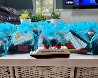 May include: A blue and white Lego cruise ship with red accents sits in front of a table filled with blue tulle bags filled with Lego bricks.
