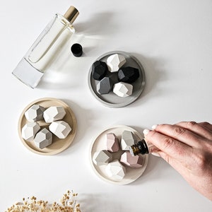 May include: Three sets of ceramic diffuser stones in grey, beige, and pink. Each set has five geometric stones in a round dish. A hand is holding a small bottle of essential oil over the pink stones.