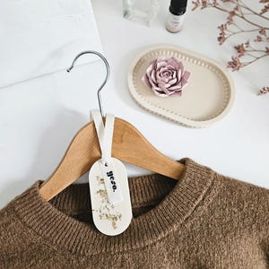 Op de afbeelding: Een bruine trui hangt aan een houten hanger met een metalen haak. Een wit label met gouden bladaccenten en het woord "yeso." is bevestigd. Een parfumfles, een flesje etherische olie en een decoratieve bloem zijn ook zichtbaar.