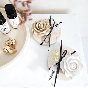 Puede incluir: Dos objetos circulares, planos y de color beige, decorados con copos de pan de oro, cada uno rematado con una rosa esculpida y atado con una cinta negra fina. Una pequeña etiqueta blanca con el texto negro yeso. está unida a cada pieza. Dos frascos pequeños y pendientes dorados están cerca.