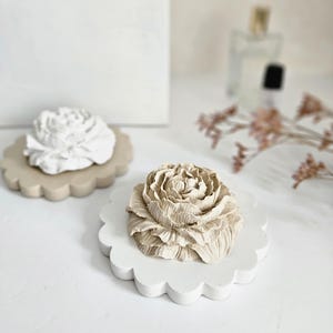 May include: Two decorative flower-shaped sculptures resting on scalloped circular bases. One sculpture is white with a beige base, and the other is beige with a white base. The flower petals feature intricate, layered textures and realistic detailing.