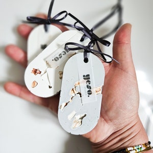 May include: Three oval-shaped, decorative tags in shades of white and gray, adorned with small, natural elements. Each tag is tied with a black ribbon. The tags have the text "yeso studio."