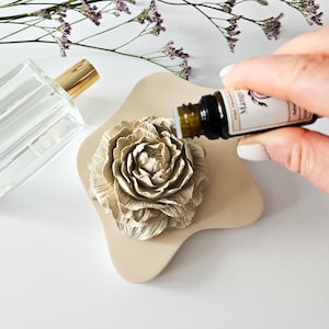 Könnte beinhalten: Ein beigefarbener, blütenförmiger Diffusor für ätherische Öle, auf den ätherisches Öl gegossen wird. Links befindet sich eine klare Glasparfümflasche mit goldenem Verschluss. Im Hintergrund lila Blüten.