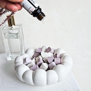 Pode incluir: Um prato branco em forma de coração cheio de ceras perfumadas em forma de coração lavanda e brancas. Óleo essencial está sendo derramado no prato. Um frasco de perfume transparente está no fundo.