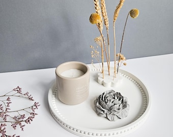 Pode incluir: Uma bandeja decorativa branca com borda de contas contém uma vela bege, uma escultura de flor cinza e trigo seco e flores amarelas. A vela tem o texto "Velvet & Vanilla". O fundo é cinza claro.
