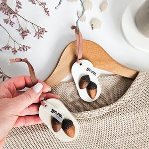 Op de afbeelding: Crèmekleurige labels met eikelontwerpen en het woord "yezo" hangen aan een houten kleerhanger. De labels zijn bevestigd met een bruin lint. De achtergrond omvat een beige trui, gedroogde bloemen en bladvormige decoraties.