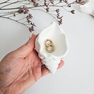 Peut inclure: Un plat en céramique blanc en forme de coquillage contient deux boucles d'oreilles créoles dorées. Le plat est tenu dans une main, avec un bracelet visible au poignet. Des fleurs violettes séchées sont en arrière-plan.