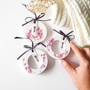 Könnte beinhalten: Drei weiße runde Ornamente mit rosa floralen Akzenten und schwarzem Band. Jedes Ornament hat ein kleines weißes Etikett mit dem Text "yeso." in Schwarz gedruckt.