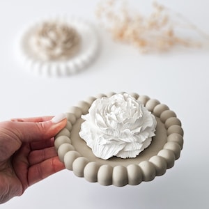 May include: A white ceramic flower with a textured surface sits on a beige ceramic dish with a beaded edge.