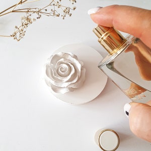 Puede incluir: Difusor de cerámica blanca en forma de rosa con tapa dorada y una botella de aceite esencial de vidrio transparente.