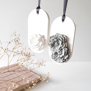 Könnte beinhalten: Zwei weiße, ovale Lufterfrischer aus Keramik mit einem schwarzen Band, das oben hängt. Ein Lufterfrischer hat ein weißes Blumendesign und der andere ein graues Blumendesign.