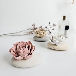 May include: Three decorative flower-shaped objects in shades of pink, gold, and white, each resting on a smooth, oval stone. The flowers are arranged on a white surface, with a sprig of dried flowers and a perfume bottle in the background.