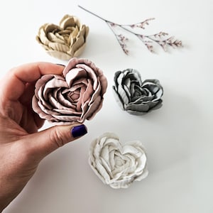 May include: Four heart-shaped ceramic flower decorations in different colours: beige, pink, grey, and white. The flowers are arranged on a white surface.