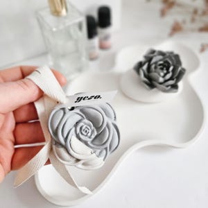 Op de afbeelding: Een grijs en wit roosvormig decoratief item met een lintlabel met de tekst "yezo". Het ligt op een witte, golvende tray. Een parfumfles en kleine flesjes etherische olie zijn zichtbaar op de achtergrond.