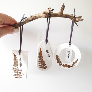 Könnte beinhalten: Drei weiße Resin-Ornamente mit getrockneten Farnblättern, die an einem natürlichen Ast hängen. Jedes Ornament hat eine andere Form: rechteckig, oval und kreisförmig. Die Ornamente sind mit dunkelvioletten Bändern aufgehängt. Jedes Ornament hat ein Etikett mit dem Text "yeso."