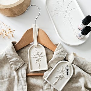 Könnte beinhalten: Ein beiges Leinenhemd hängt an einem Holzkleiderbügel, verziert mit zwei cremefarbenen Keramikanhängern mit Palmenmuster. Ein passendes Tablett und zwei kleine ätherische Ölflaschen sind ebenfalls zu sehen, zusammen mit einem Holzkerzenhalter.