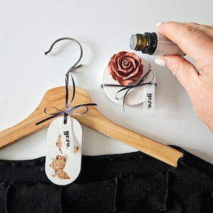 Può includere: Un appendiabiti in legno con un'etichetta in ceramica bianca che dice "osoli" e un diffusore di rose in ceramica marrone con un'etichetta bianca che dice "rosa". Una mano tiene una piccola bottiglia di olio essenziale sopra il diffusore di rose.