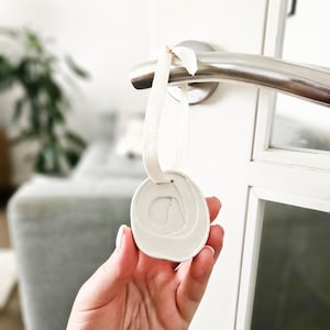 Könnte beinhalten: Ein runder, cremefarbener Türanhänger aus Keramik mit strukturierter Oberfläche und cremefarbenem Band. Der Türanhänger hängt an einem silbernen Türgriff. Der Hintergrund ist verschwommen und zeigt eine weiße Tür und einen Blick in ein Wohnzimmer.