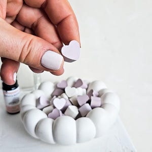 Op de afbeelding: Een witte keramische kom gevuld met hartvormige snoepjes in tinten wit en lavendel. Een hand houdt een lavendelkleurig hartvormig snoepje boven de kom. De kom staat op een wit oppervlak.