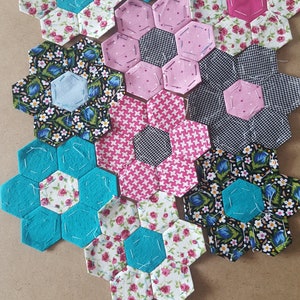 Könnte beinhalten: Neun Stoff-Sechsecke in verschiedenen Blumenmustern, darunter rosa, blau und schwarz-weiß, mit türkisfarbenen Akzenten. Die Sechsecke sind in einer Gruppe auf einer braunen Oberfläche angeordnet.