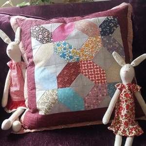 Könnte beinhalten: Zwei weiße Stoffhasenpuppen mit rot-weißen Blumenkleidern sitzen auf einem lila Samtsofa mit einem Patchwork-Kissen. Das Kissen ist aus verschiedenen Stoffen in den Farben Rosa, Rot, Blau und Braun.