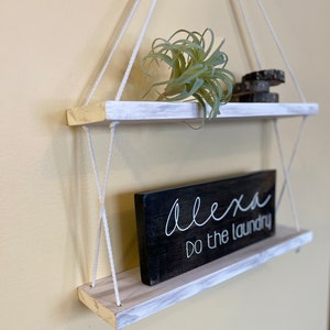 May include: White painted wooden shelves with rope hanging system. The shelves are decorated with a small green plant and a black sign that reads "Alexa do the laundry".