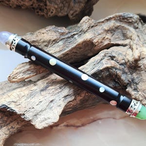 May include: A dark wooden wand with white dot inlays and silver accents. The wand has a light purple crystal on one end and a green crystal on the other. The wand is resting on a piece of driftwood.
