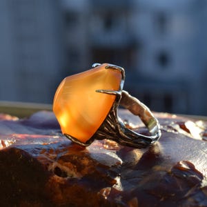 May include: A silver ring featuring a large, orange-hued gemstone. The gemstone is rectangular with rounded edges, set in a textured, branch-like silver band. The ring is displayed on a textured, dark surface.