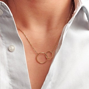 Peut inclure: Un collier doré avec deux cercles entrelacés. Le collier est présenté sur une chemise boutonnée à rayures bleu clair et blanches. Les cercles sont reliés par une chaîne délicate.
