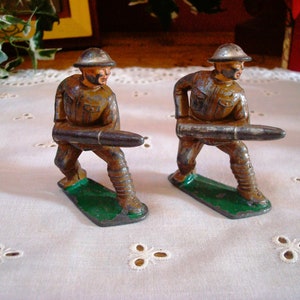 May include: Two toy soldiers made of metal, painted brown and green, are holding large guns. The soldiers are standing on a white surface.