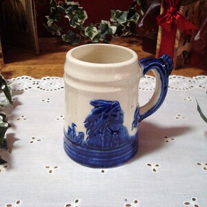 May include: A white ceramic beer stein with a blue and white floral design. The stein has a handle and a wide base.