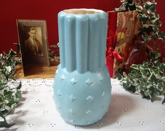May include: A light blue ceramic vase with a textured, diamond-shaped pattern. The vase has a cylindrical shape with a slightly flared top.