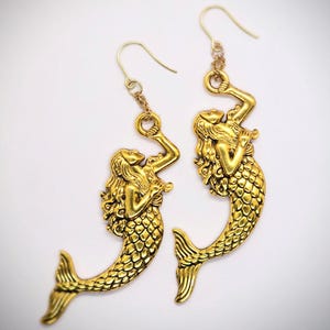 May include: A pair of gold mermaid dangle earrings. Each earring features a detailed mermaid design with a scaly tail and flowing hair. The mermaids are holding onto a ring at the top of the earring.