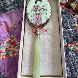 May include: An antique hand mirror with a jade handle and an oval-shaped mirror. The mirror features a painted image of a woman in traditional attire. The mirror is in a white box with a red string attached.