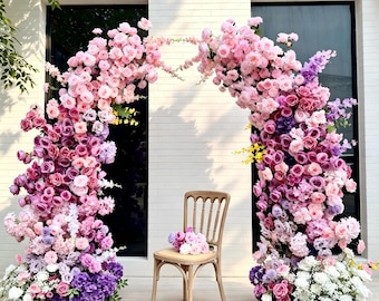 May include: A large floral archway composed of numerous artificial roses and hydrangeas in shades of pink, purple, and white, arranged in a dense, cascading display. A light-colored wooden chair with a slatted back sits centered beneath the arch.