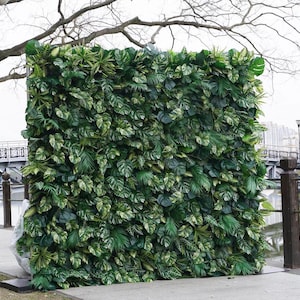 May include: A large, rectangular artificial green wall composed of various types of lush, vibrant green leaves. The leaves have different shapes and textures, creating a dense, natural-looking display. The wall is set outdoors, with a bridge and trees in the background.