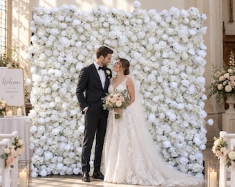 May include: A large, rectangular wedding flower wall backdrop composed of dense, white artificial roses and hydrangeas. The wall serves as a decorative display for a wedding ceremony, featuring a uniform arrangement of white floral blooms.