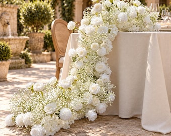 May include: A long floral table runner composed of white roses and baby's breath flowers, cascading from the top of a beige tablecloth down to the ground, creating a continuous arrangement of white blossoms and delicate greenery.