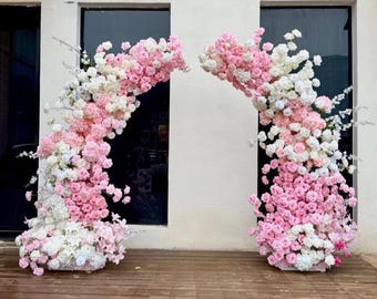 Puede incluir: Dos grandes arreglos florales curvos que presentan una densa mezcla de rosas blancas y de varios tonos de rosa, junto con flores más pequeñas. Estas estructuras decorativas están diseñadas para mantenerse en pie, creando un arco simétrico.