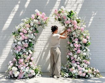 May include: A large, arched floral display featuring a dense arrangement of pink and white roses, hydrangeas, and greenery. The structure is composed of two separate sections forming a curved frame, decorated with various blossoms and foliage.