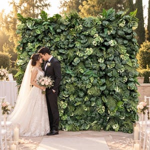May include: A large, rectangular artificial green wall backdrop densely covered with various tropical leaves, including monstera and fern foliage in shades of dark and light green, standing outdoors as a decorative feature for a wedding ceremony.