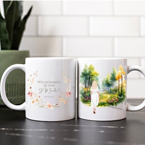 Könnte beinhalten: Zwei weiße Keramikbecher mit einem Blumendesign und einer Aquarellillustration einer Frau, die durch einen Wald geht. Ein Becher sagt "Willkommen in der JW Familie!" mit einem Blumengekränz um den Text.