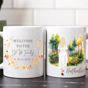 Könnte beinhalten: Zwei weiße Keramikbecher mit Blumenmuster. Ein Becher sagt "Willkommen in der J W Familie! 02.11.2024" und der andere Becher hat eine Aquarellillustration einer Frau, die in einem Wald spaziert, mit dem Namen "Nathalie" darunter.