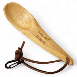 Puede incluir: Una cuchara de madera con una correa de cuero y un mosquetón. La cuchara está grabada con "Nordic Wood Germany".