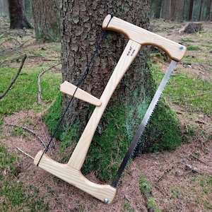Puede incluir: Una sierra de arco de madera con un mango de cordón negro y una hoja plateada. La sierra está apoyada en un lecho de agujas de pino y musgo.